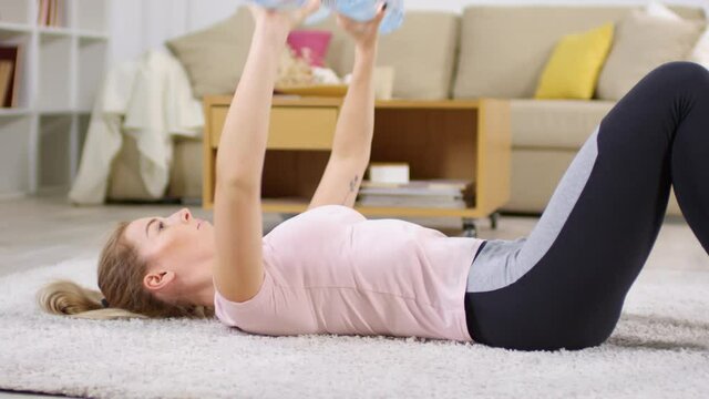 Young woman in sportswear lying on carpet at home and doing dumbbell floor press with water bottles during workout