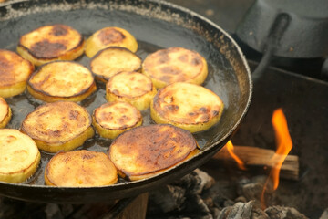 Man is frying a slices of fresh organic zucchini on frying pan with hot boiling oil on coals in grill, hand closeup. Preparing food in chargrill on nature. Burning flame is under the pan.
