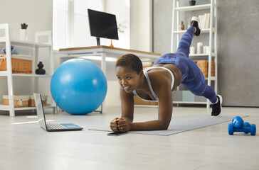 Fototapeta na wymiar Young african american woman doing sports exercises in front of laptop during online broadcast of fitness workout. Woman trains on a sports mat at home in the living room. Concept of active lifestyle.