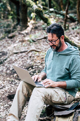 Obraz premium Adult caucasian man sitting on the ground in the forest do a online work connection with laptop computer - internet wireless smart working job digital nomad remote worker lifestyle