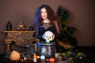 Portrait of a witch with a skull in hands standing next to a cauldron and a table with herbs and ingredients for witchcraft.