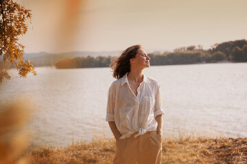 Beautiful young relaxed woman in white blouse enjoying nature breathing fresh air meditating on the river on an autumn day - obrazy, fototapety, plakaty