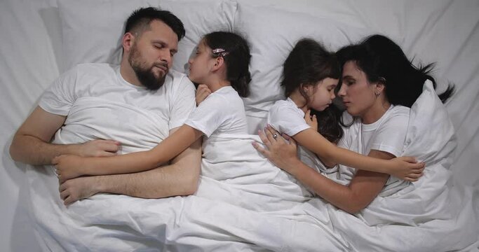 Top view of Caucasian family with two children sleeping in bed with white sheets. High angle shooting. A family of four is sleeping. Mom, dad and daughters good night, restful sleep 4k