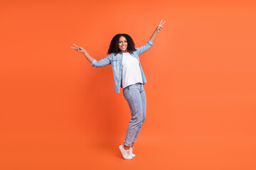 Fototapeta na wymiar Full body photo of cool young brunette lady show v-sign wear jeans shirt shoes isolated on orange background