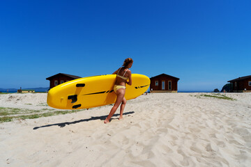 Back view relaxed female surfer with tanned skin in yellow swimsuit standing on beach with surfboard
