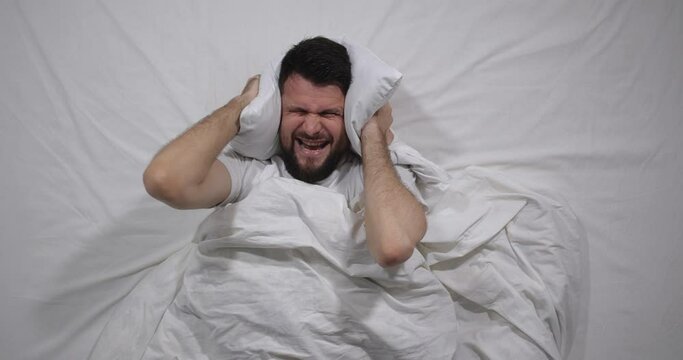  Top view  of caucasian bearded man lying awake  in bed with white linens in light room covering  ears with a pillow. Attractive guy suffers from loud noise from neighbors behind the wall  4K 