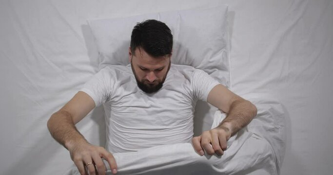  Top view of handsome bearded guy lying in bed with white linens in light room. High angle shooting. Man is looking down under the blanket and showing, thumbs up. Male health concept