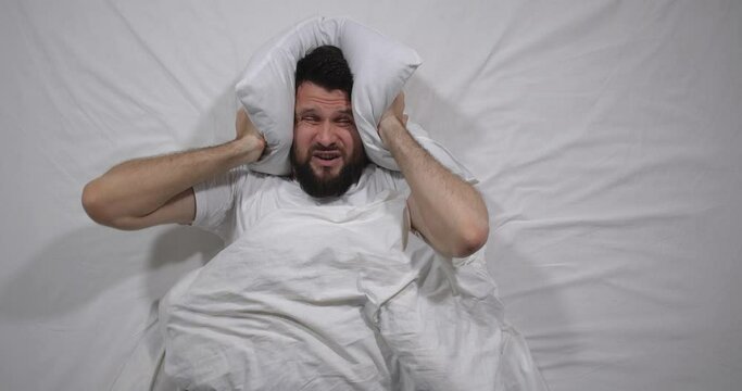  Top view  of caucasian bearded man lying awake  in bed with white linens in light room covering  ears with a pillow. Attractive guy suffers from loud noise from neighbors behind the wall  4K 