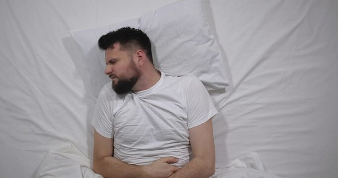 Top view  of caucasian bearded man lying in bed with white linens in light room suffering from stomach ache. Attractive guy suffers from pain migraine holding stomach with hands   4K 
