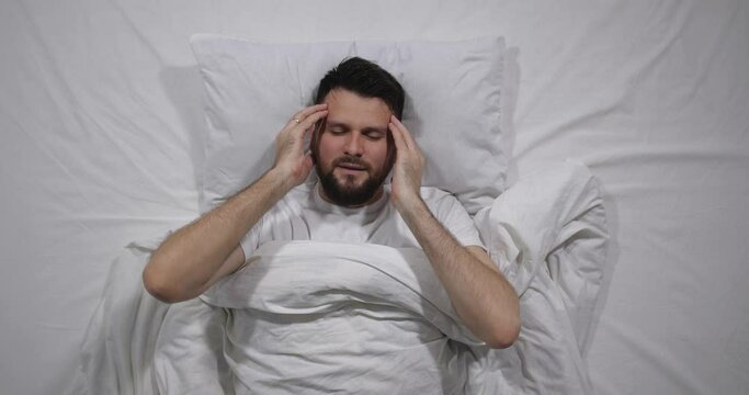  Top view  of caucasian bearded man lying awake  in bed with white linens in light room covering  ears with a pillow. Attractive guy suffers from loud noise from neighbors behind the wall  4K 