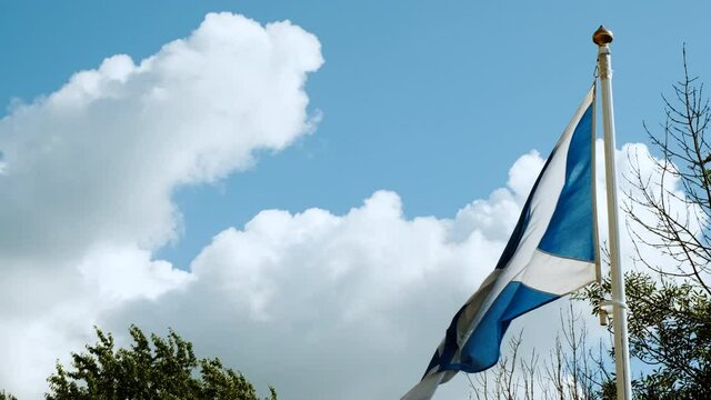 Slow motion view of the flag of Scotland, also known as St Andrews Cross, flying against a blue sky background with white clouds