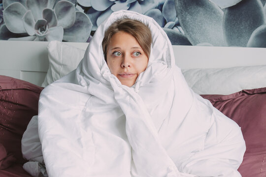A young frozen funny caucasian charming blonde woman hides under a large white warm blanket to escape from the cold sitting on the bed in her room. The problem of heating in the house