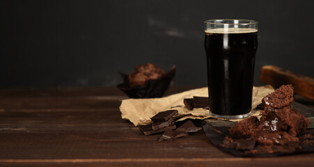 Glass of beer stout standing on wooden board next to chocolate muffin. Horizontally stretched panoramic image for banner - obrazy, fototapety, plakaty