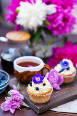 Cupcake with cream decorated with violet flowers lying next to a mug of tea among the flowers of peonies and scattered honeysuckle berries
