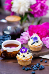 Cupcake with cream decorated with violet flowers lying next to a mug of tea among the flowers of peonies and scattered honeysuckle berries