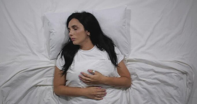  Top view  of caucasian woman lying in bed with white linens in light room suffering from toothache. Attractive brunette suffers from dental  pain  holding face with hands   4K 