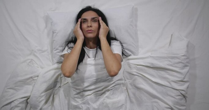  Top view  of caucasian woman lying in bed with white linens in light room suffering from headache. Attractive brunette suffers from pain migraine holding head with hands   4K 