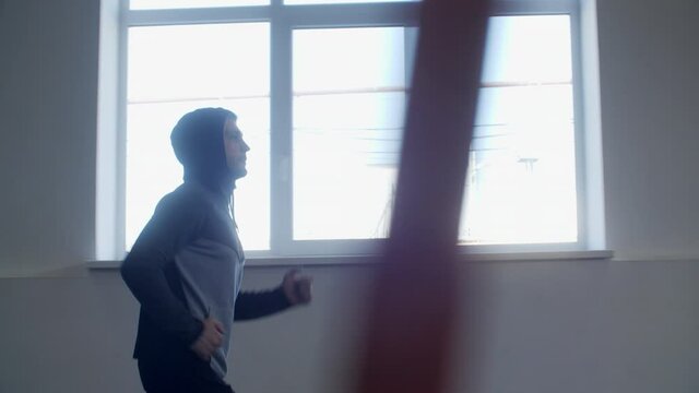 Side view of athletic man in hood running indoors. Muscular focused man in sportswear jogging during fitness workout