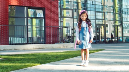 Adorable beautiful Caucasian little girl smiling in front of school on sunny day. Pretty cute school kid standing with smile on face outdoor on street with pink backpack. Back to school. Study concept