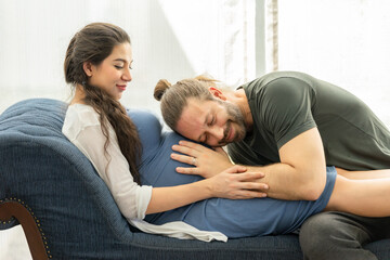 Smiling father leaning to pregnant wife and listening to his wife's tummy on a sofa by sunny windows in apartment. Multi-Ethnic couple spending time together at home. Healthcare, Pregnancy, Motherhood
