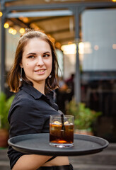 Portrait young waitress standing in outdoor cafe. girl the waiter holds a tray with a glass of alcoholic cocktail