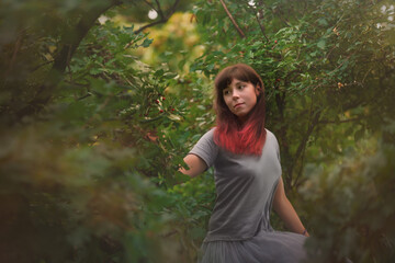 A young woman with brownish-red hair in a grey shirt and skirt is sitting on a tree, touching a branch with red berries.