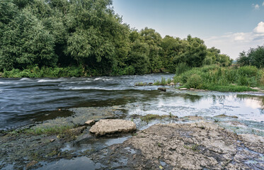 A river with patches of grass and stones of scattered running to the horizon. Long exposure shot. The banks overgrown with lush vegetation. Generic vegetation, scenic destinations, landscapes.