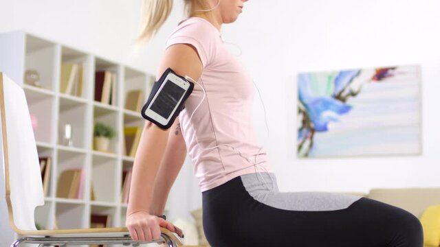 Tilt up shot of young caucasian woman in sportswear listening to Music on smartphone in armband and doing triceps dips with chair while training at home