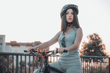 Muchacha joven parada y aparcada con una bicicleta y casco en la ciudad junto a los arboles y la naturaleza de un parque - obrazy, fototapety, plakaty