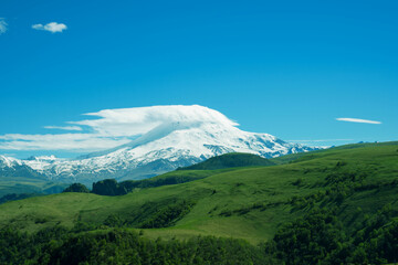 Snowy peak of Elbrus mount covered by clouds among green mountain landscape