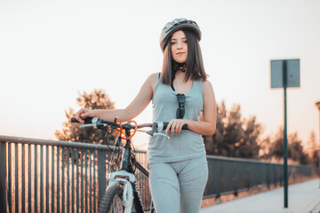 Joven adolescente parada en la ciudad junto con una bicicleta y casco haciendo deporte - obrazy, fototapety, plakaty