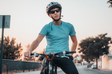 Muchacho joven con casco y gafas de ciclista haciendo ejercicio con la bicicleta junto a la puesta de sol en la ciudad