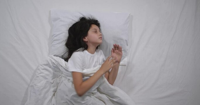  Top view  of girl 9 years old with long brown waking up in bed with white linens in light room. High angle shooting. Teenager girl getting out of bed. Good morning calm awakening 4k 