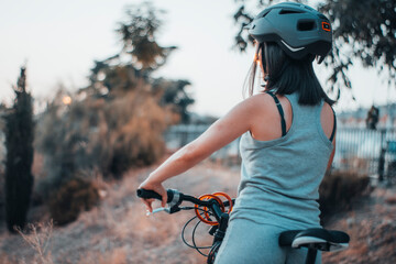 Chica joven de espaldas haciendo ciclismo y ejercicio en un parque junto a los arboles con un casco...