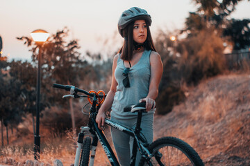 Mujer joven con un casco de protección de pie parada con una bicicleta haciendo ejercicio en un parque - obrazy, fototapety, plakaty