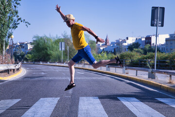 Hispanic mature adult man with hat, mustard-colored t-shirt and short jeans making fun jumps over a crosswalk in the middle of the city.