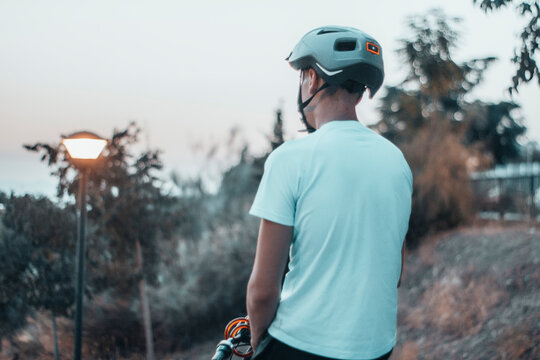 Hombre joven paseando con una bicicleta de espaldas con un casco de protecci&oacute;n en un parque al anochecer