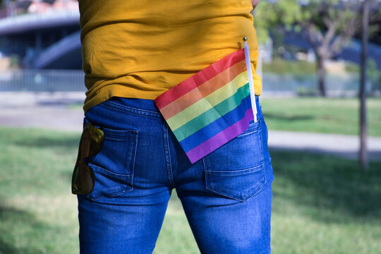 Mature adult man in a mustard-colored T-shirt and short jeans with sunglasses in one back pocket and gay pride flag in the other.