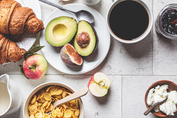 Breakfast table with cornflakes, croissants, fruit, cheese and cup of coffee. Gray tile background, top view.