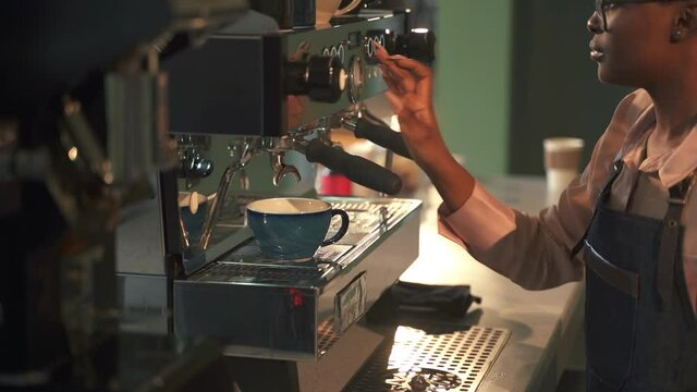 Barista Afro-American woman works on coffeemaker. Spbas Black lady puts blue ceramic cup on rack under small taps at counter and press button in coffee shop