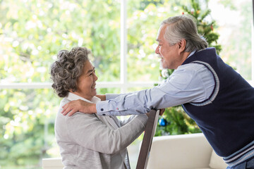 Obraz premium Happy Asian senior man embracing his wife on Christmas day at home. Senior couple spending time together with Christmas tree. Merry Christmas and Happy Holidays. Christmas holiday celebration