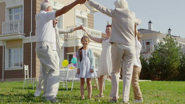 Happy Multigenerational Family In Party Hats Holding Hands And Dancing Around Little Birthday Girl While Having Celebration Outdoors In The Backyard On Sunny Day