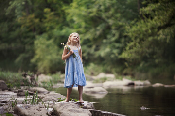 a little girl in a blue sundress with a bouquet of daisies walks by the river splashes of water