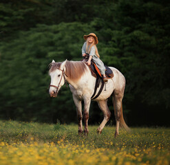 a little girl in a blue linen dress and a straw hat is stroking a horse riding