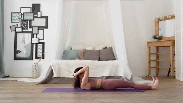 Slender, flexible, caucasian middle-aged woman with black hair in beige tracksuit performs standing relax asanas Fish Pose, Matsyasana on purple yoga mat in bright room with bed, a mirror, a window