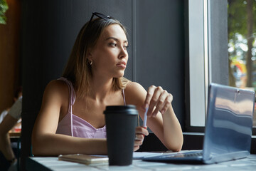 beautiful young asian woman sitting at table near window, working on laptop. beautiful lady studying, doing homework, having cup of coffee. modern online education, distant work, remote job, freelance