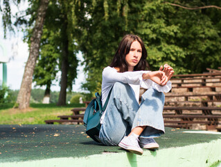 Naklejka premium A teenage girl with a bored face sits with a backpack on the sidewalk and looks into the distance