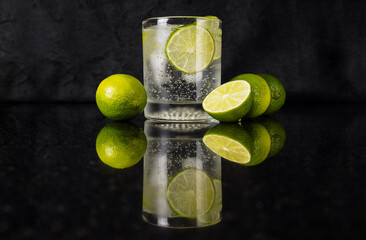 Sparkling water with slice of lime and limes on reflected black surface with black background