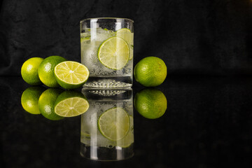 Sparkling water with slice of lime and limes on reflected black surface with black background