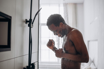 Relaxed young African-American man takes shower standing under hot water jets in contemporary unit...